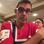 A man wearing glasses and a red patterned shirt looks at the camera, with a woman visible out of focus behind him